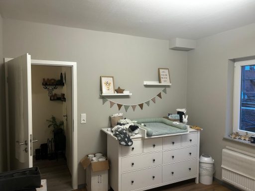 Heller Raum mit Wickeltisch, Regalen und offener Tür zu einem weiteren Raum.